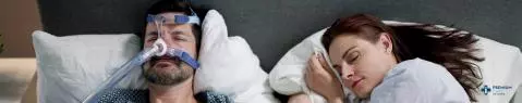 Un homme portant un masque PPC est allongé dans le lit, adossé à un oreiller, tandis qu'une femme à côté de lui sourit et regarde une tablette qu'ils tiennent ensemble.