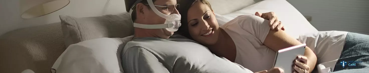 Un homme portant un masque PPC est allongé dans le lit, adossé à un oreiller, tandis qu'une femme à côté de lui sourit et regarde une tablette qu'ils tiennent ensemble.